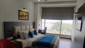 A hotel room with a large bed, a desk, a chair, a small table, and a window with a view. - JB SKY HILTON INN (Lucknow)