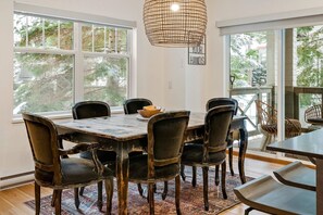 A dining area with a large table and chairs, large windows with a view of trees, and a hanging light fixture.