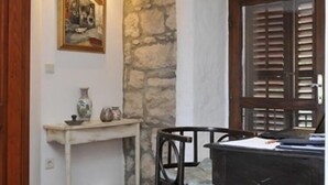 Quarto com parede de pedra, porta de madeira com persianas, escrivaninha com cadeira, mesa pequena com objetos decorativos e quadro na parede.