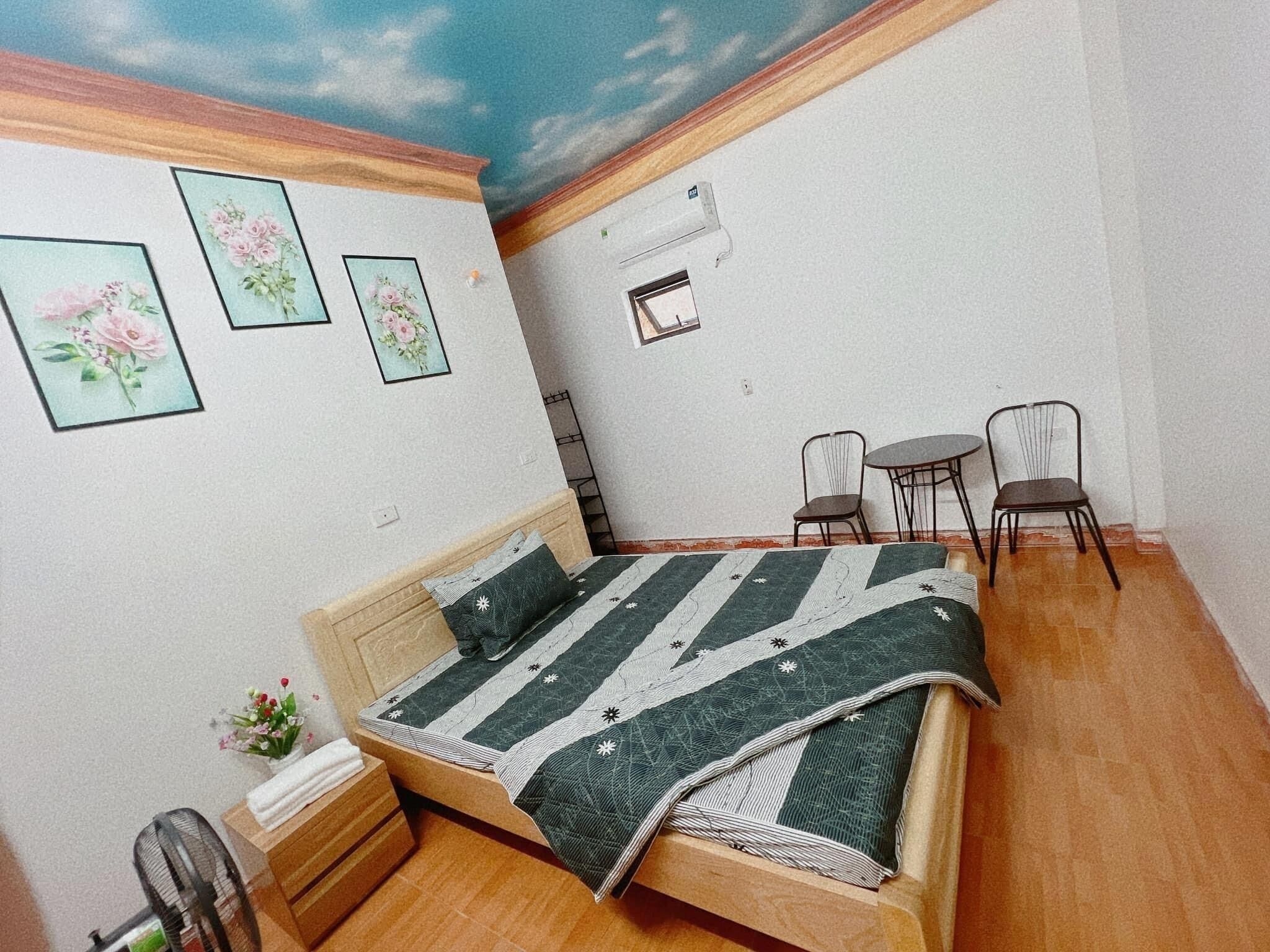 A bedroom with a bed, two chairs, a small table, a fan, and a ceiling mural.