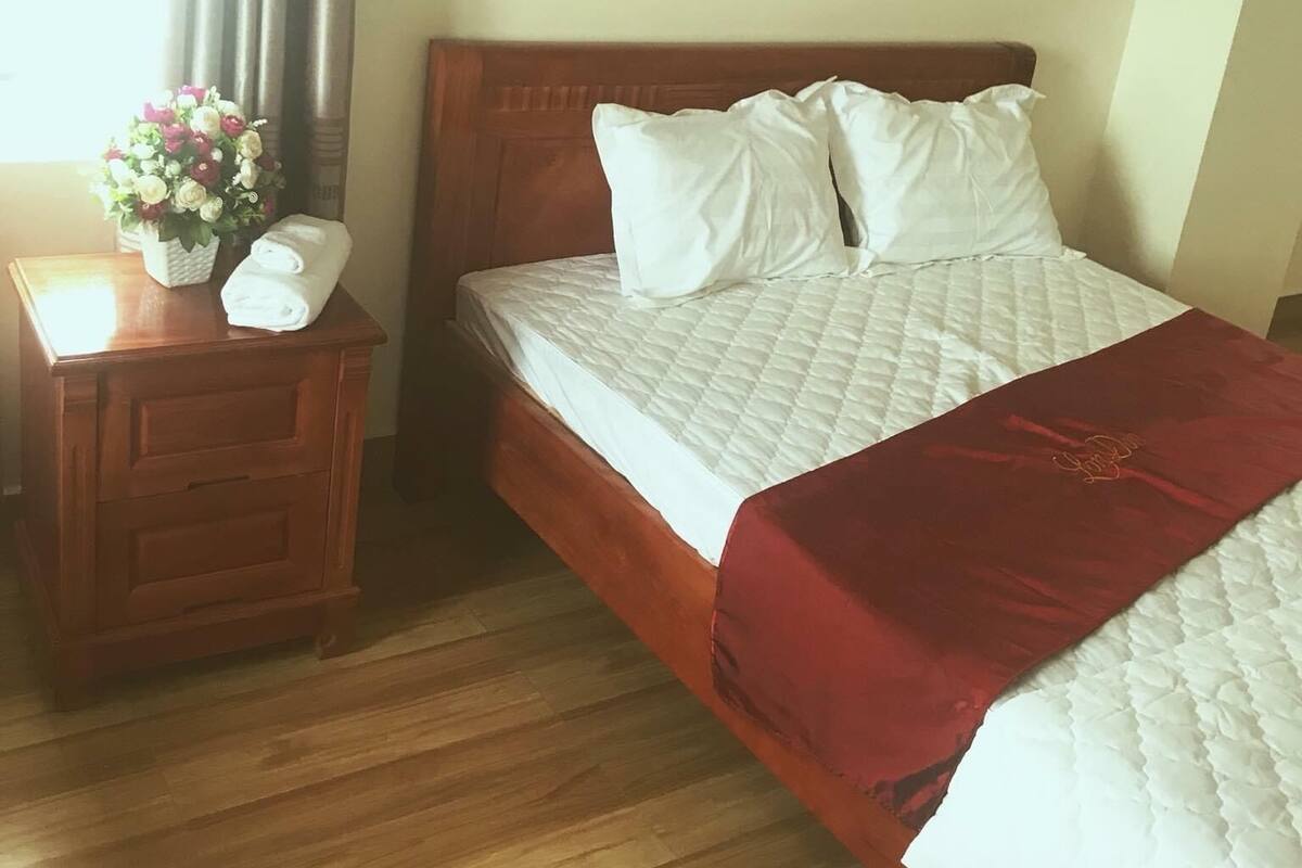 Una cama bien hecha con ropa blanca y una manta roja, una mesita de noche de madera con un florero y un piso de madera.