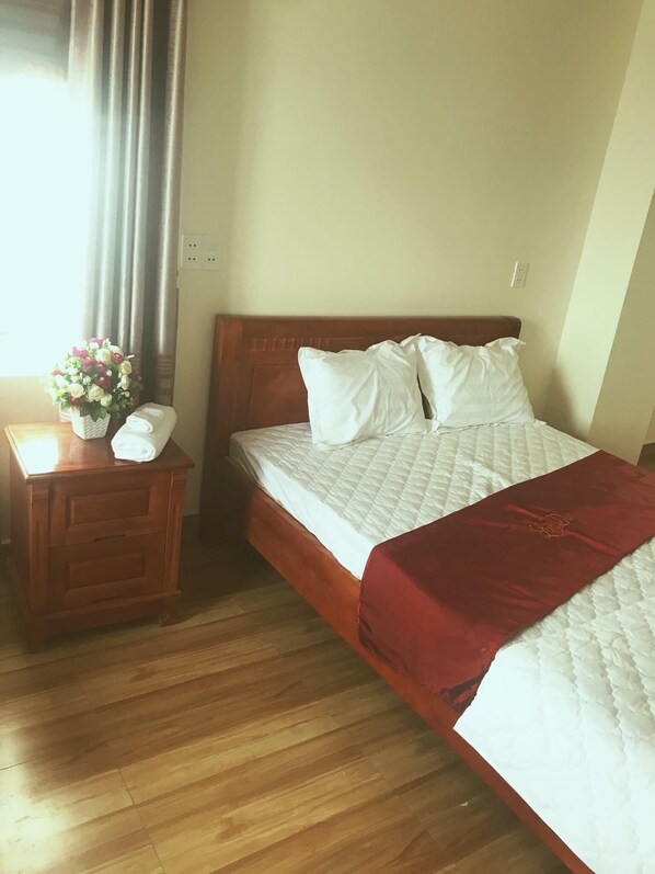 A neatly made bed with white bedding and a red blanket, a wooden nightstand with a vase of flowers, and a wooden floor.