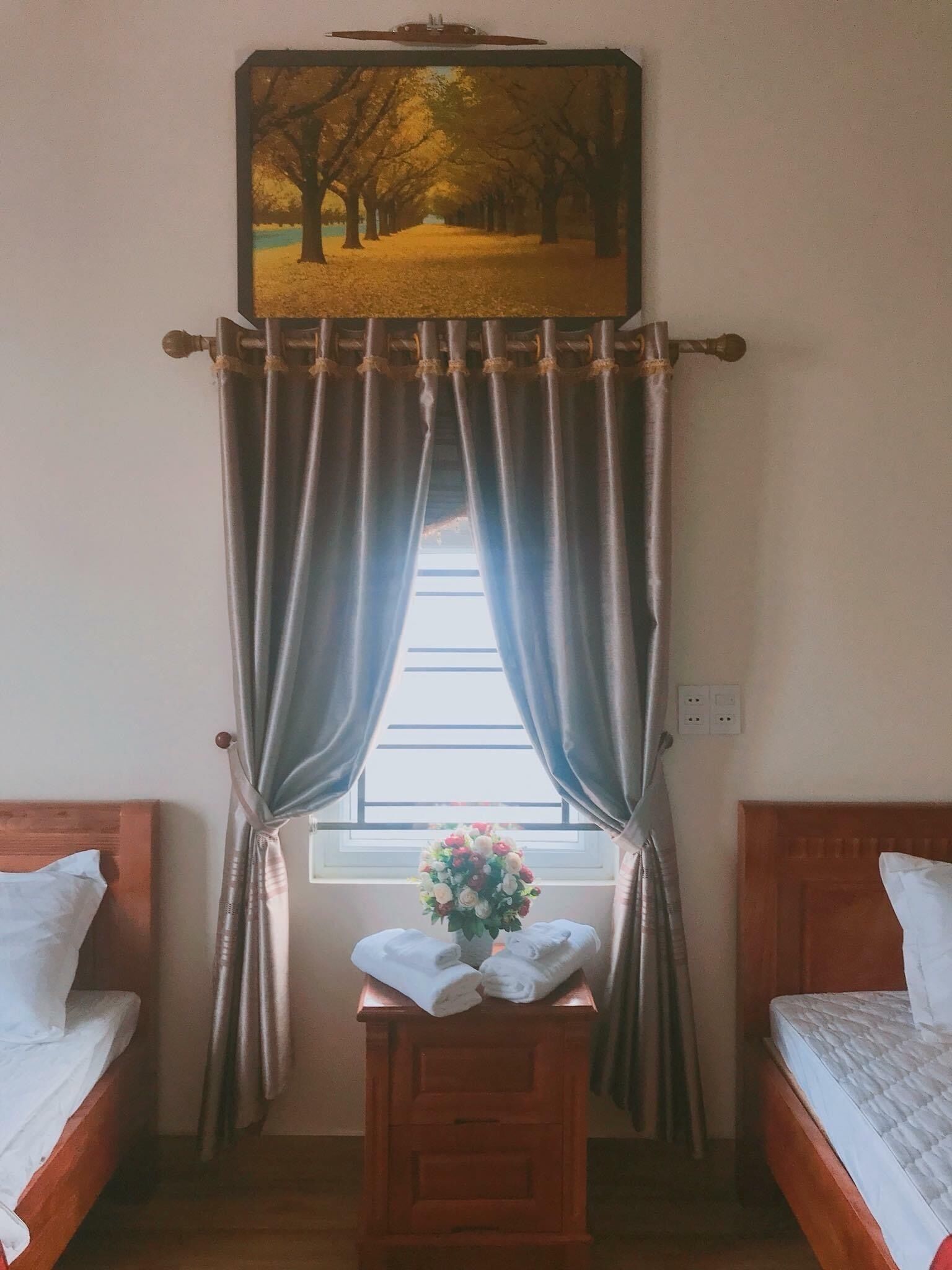 Un dormitorio con dos camas individuales, una mesita de noche de madera con un jarrón de flores y un cuadro de un camino bordeado de árboles sobre la ventana.