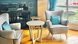 A modern living room with a piano, two armchairs, a small round table, and a large window with a view of buildings.