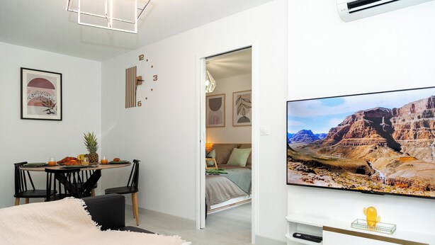 A modern living room with a flat-screen TV, a dining area with a table and chairs, and a cozy seating area with a sofa and a blanket.
