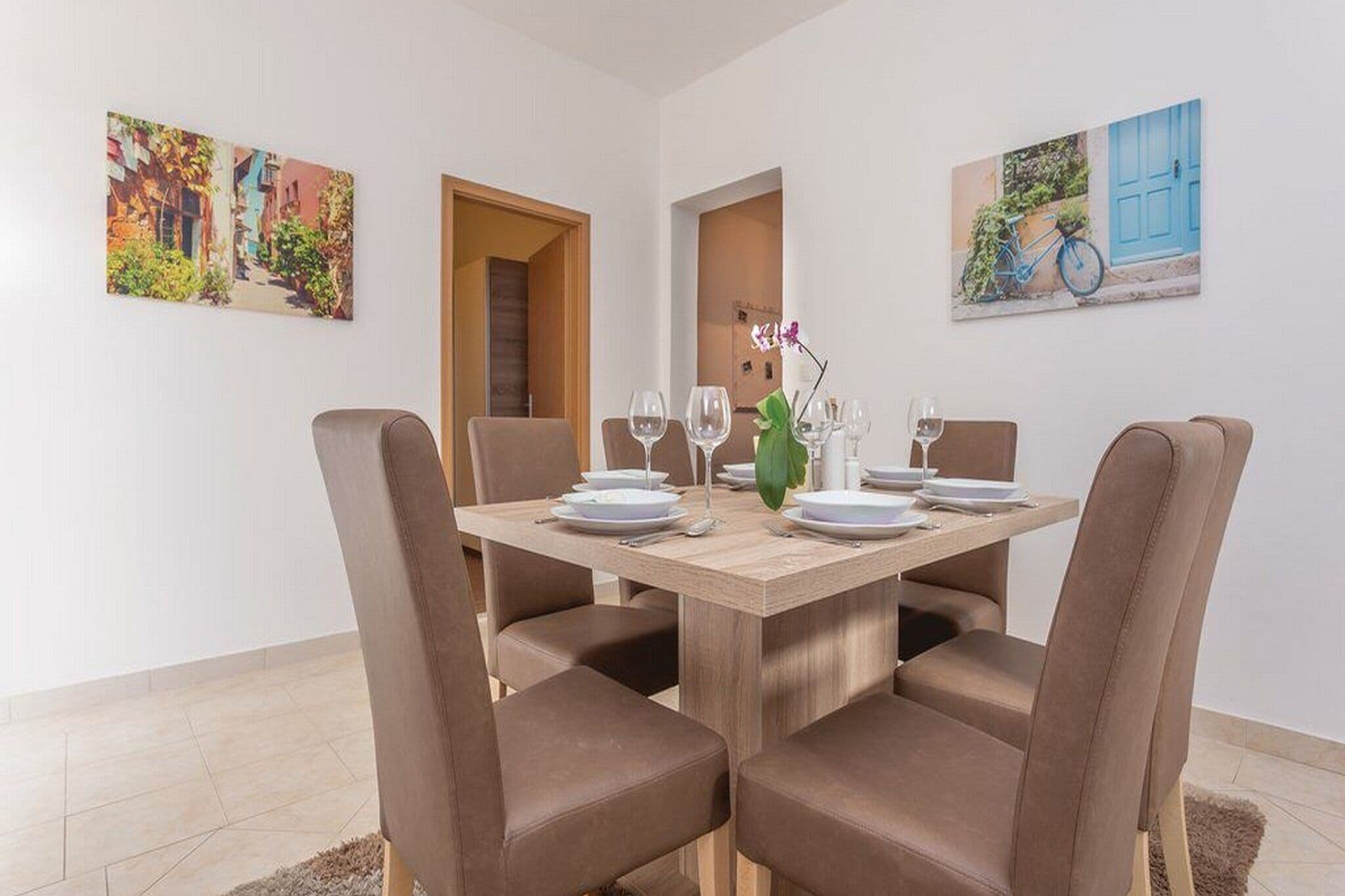 A dining area with a wooden table set for four, surrounded by brown chairs. There are two paintings on the walls, one depicting a street scene and the other a blue door with a bicycle.