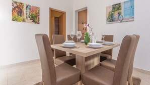 Ein Essbereich mit einem Holztisch, gedeckt für vier Personen, umgeben von braunen Stühlen. An den Wänden hängen zwei Gemälde, eines zeigt eine Straßenszene, das andere eine blaue Tür mit einem Fahrrad.
