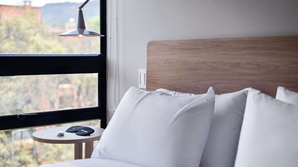 A neatly made bed with white linens and pillows, a small round table with a lamp, and a view of a mountainous landscape through a large window. - w* | Brilliant Loft near Parque 93 (Bogotá)