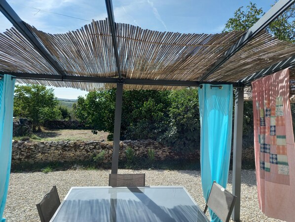 A covered outdoor seating area with a table and chairs, a stone wall, and a view of a garden and hills in the background.