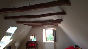A small, cozy attic bedroom with a sloped ceiling, a single bed with a red pillow, a chair, a small table with a vase, and a window with a view of greenery. - Apartment with garden and pool near fabulous sights (Rocamadour)