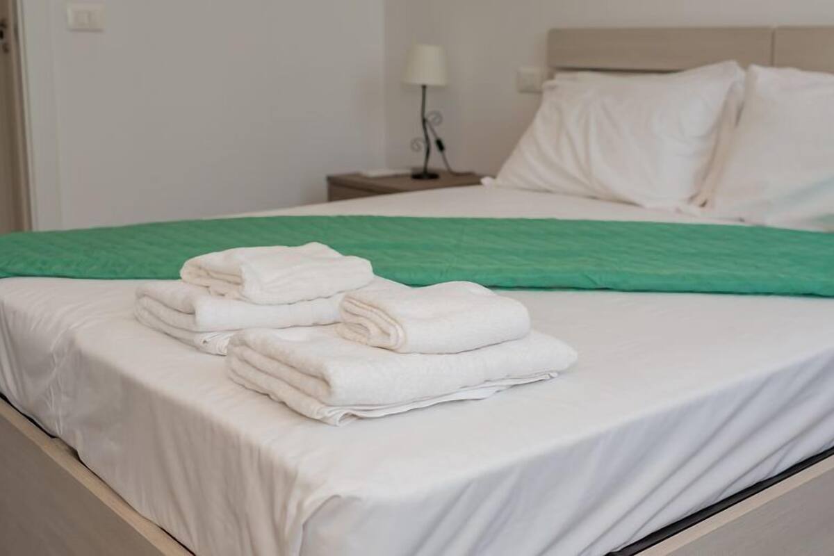A neatly made bed with white linens and a green throw blanket, a bedside lamp, and a white pillow on a wooden headboard.