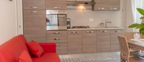 Una cucina compatta con elettrodomestici moderni, una zona pranzo con tavolo e sedie, e un divano rosso.