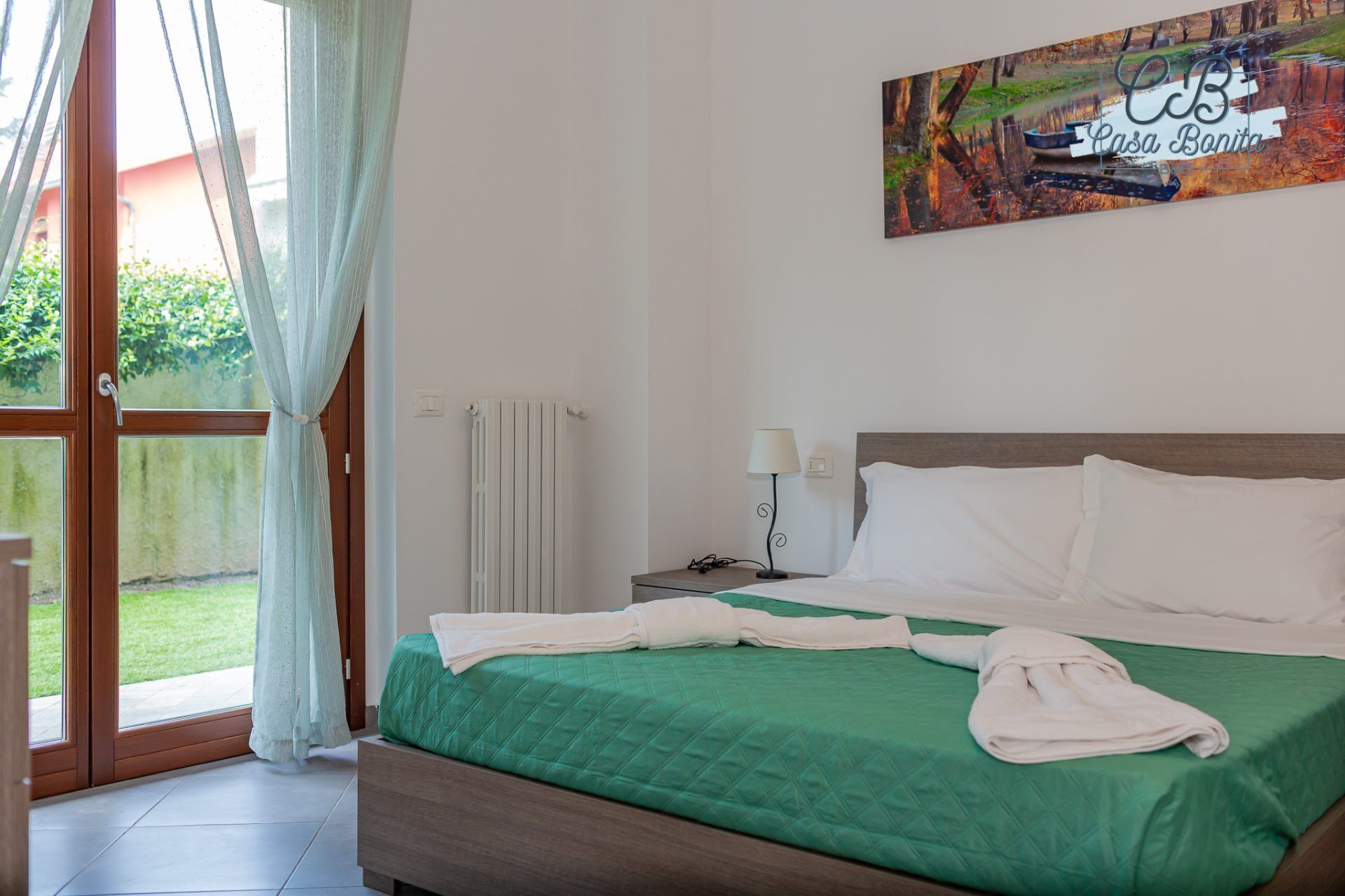 Un dormitorio con colcha verde, almohadas blancas y una mesita de noche con lámpara.