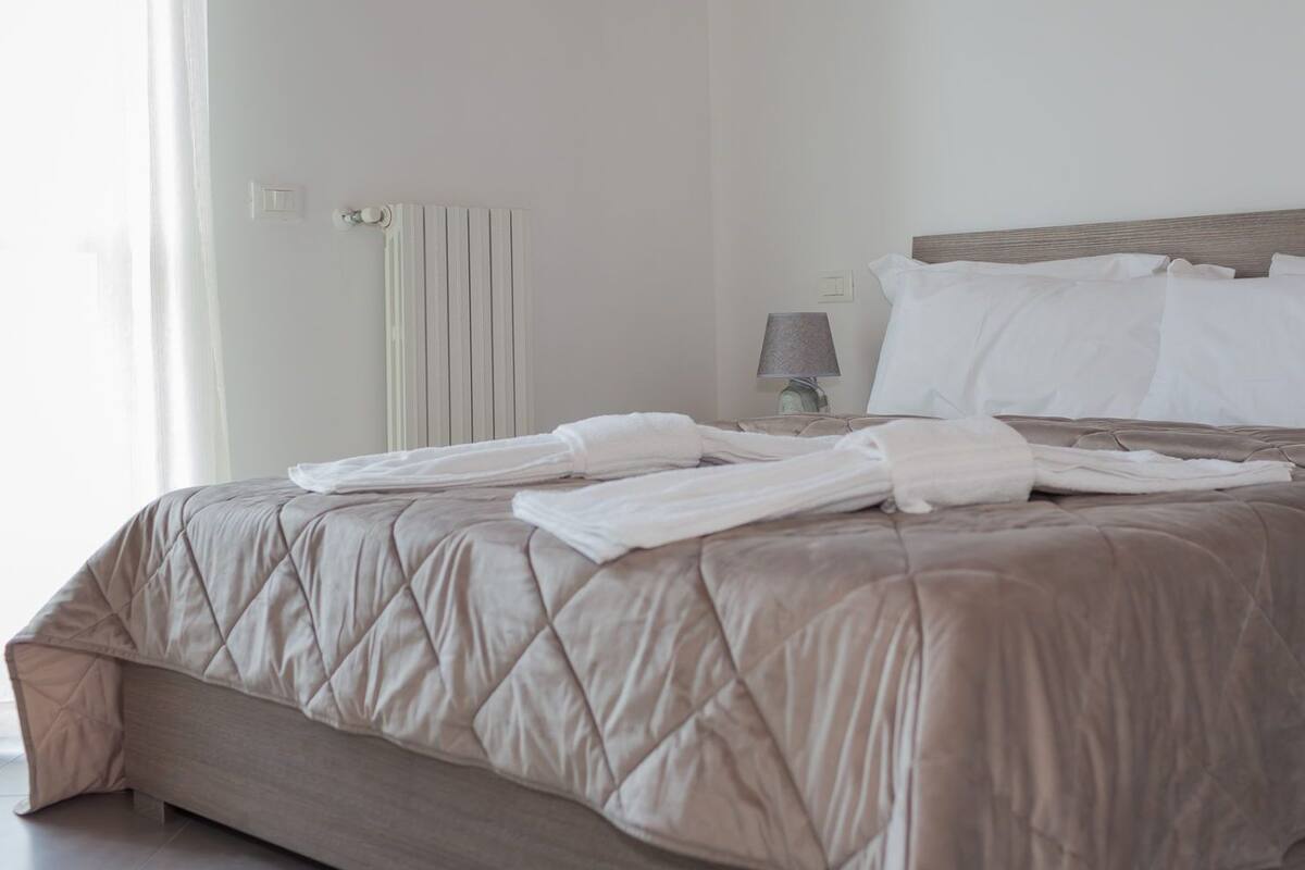 A neatly made bed with a beige quilted bedspread and white pillows, a bedside lamp, and a radiator on the wall.