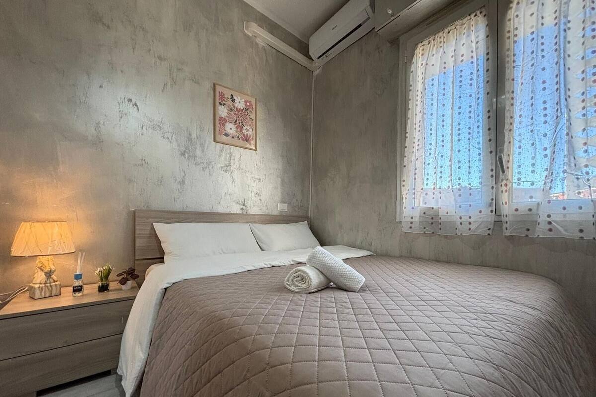A bedroom with a quilted bedspread, a bedside table with a lamp, and a window with sheer curtains.