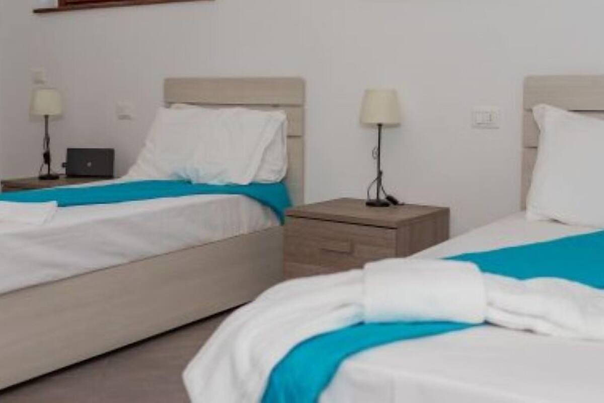 A hotel room with two beds, each with a white and blue bedspread, a wooden nightstand with a lamp, and a laptop on one of the beds.