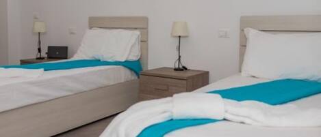 Ein Hotelzimmer mit zwei Betten, jedes mit einer weiß-blauen Bettdecke, einem Nachttisch aus Holz mit einer Lampe und einem Laptop auf einem der Betten.