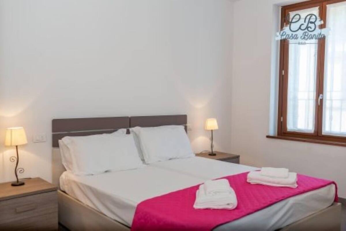 A single bed with white linens, a pink blanket, and two pillows. There are two bedside tables with lamps.