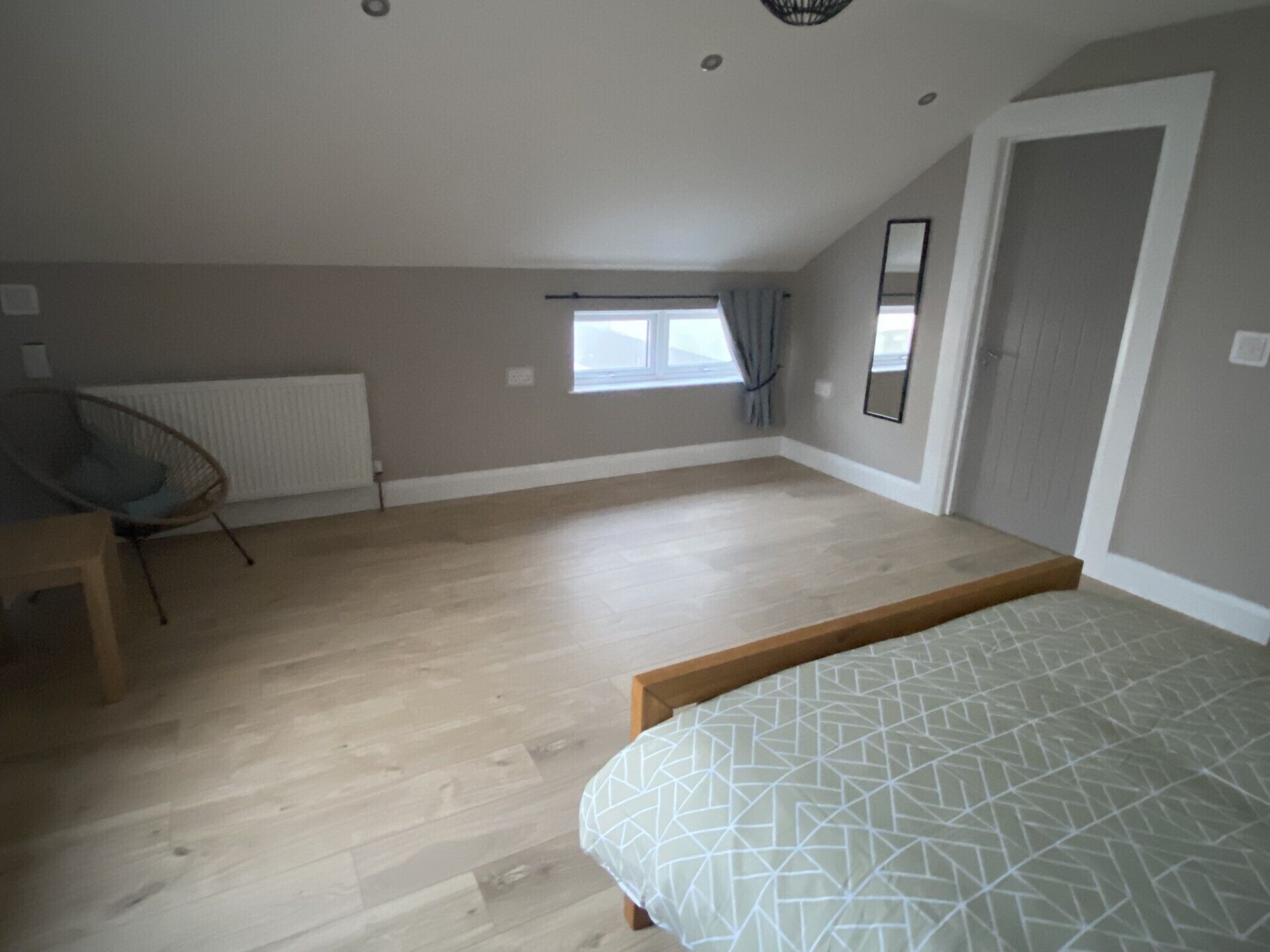 A spacious room with a sloped ceiling, a large window with curtains, a radiator, and a bed with a geometric-patterned bedspread.