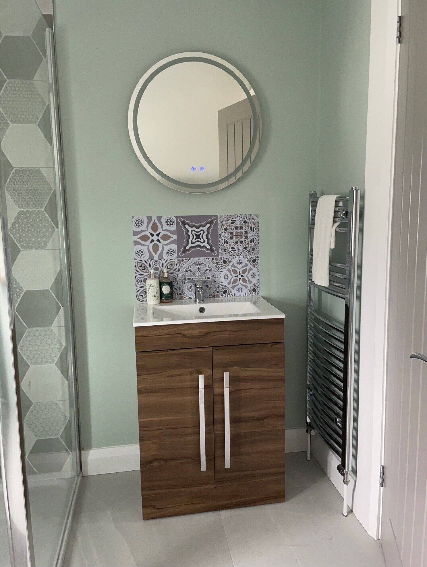 Une salle de bain avec un meuble-lavabo en bois, un miroir rond, un pare-chocs en carreaux à motifs et un porte-serviettes.