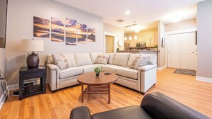A living room with a sofa, coffee table, and a TV stand. There is a kitchen in the background with cabinets and a microwave. - 6BR Downtown | Side-by-Side Townhomes | 2 Garages (Indianapolis)
