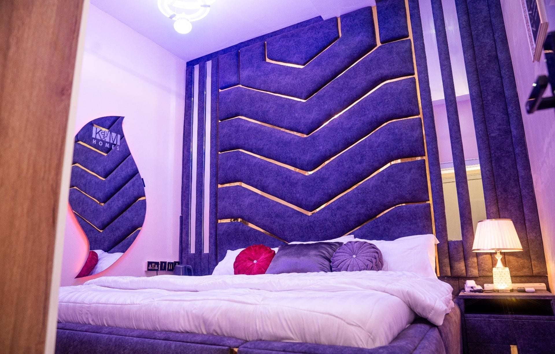 A modern bedroom with a large bed, a decorative headboard, a bedside table with a lamp, and a mirror.