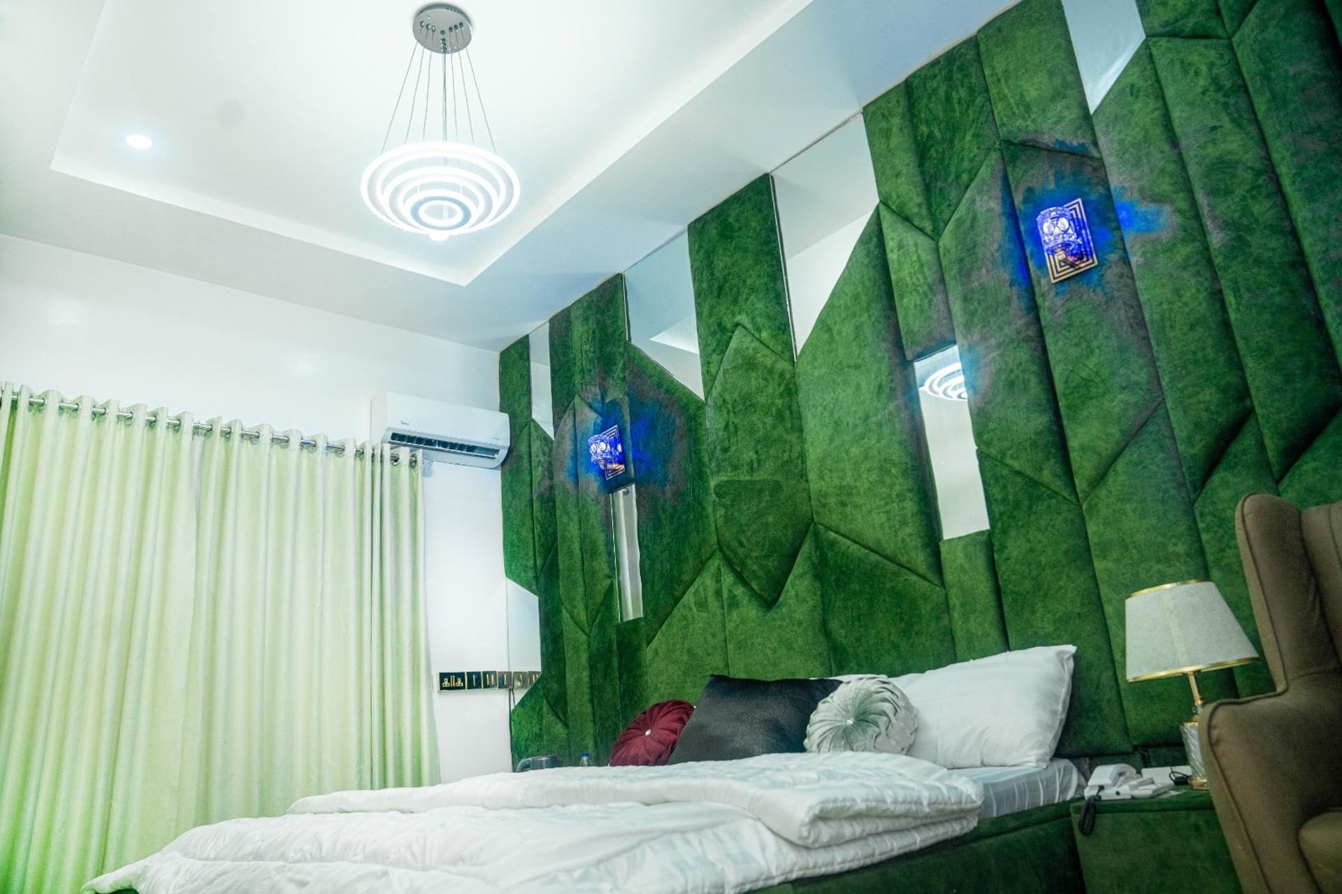 Un dormitorio moderno con una pared verde texturizada, una cama con ropa blanca, una mesita de noche, una lámpara y un equipo de aire acondicionado.