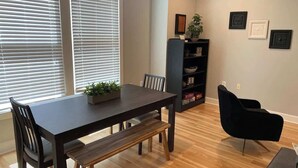 Uma área de refeições com uma mesa de madeira escura, bancos e cadeiras. Há uma cadeira preta e uma estante com objetos decorativos na parede.