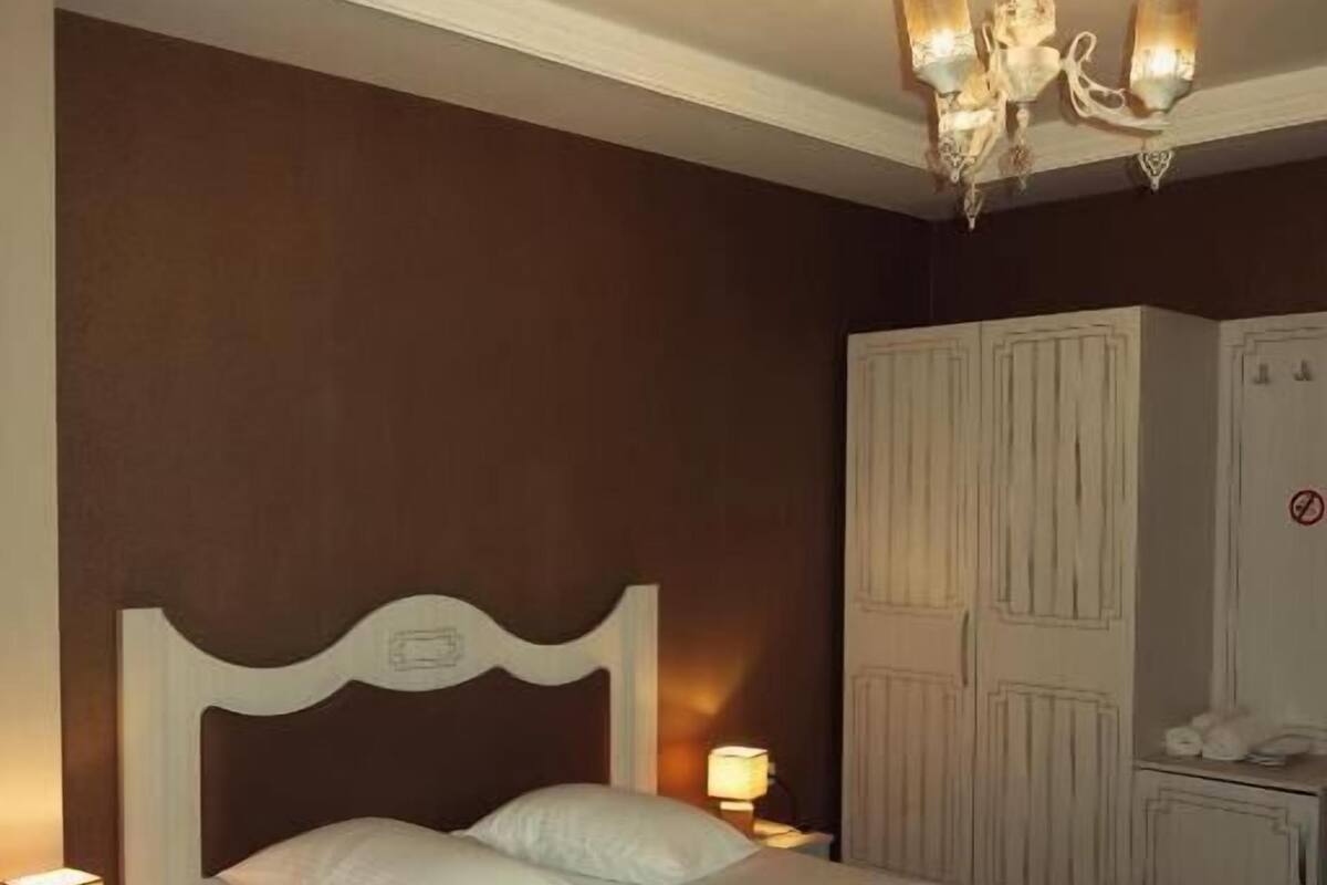 A neatly made bed with white linens, a wooden headboard, and a chandelier above. There is a wardrobe with a mirror and a telephone on the right.