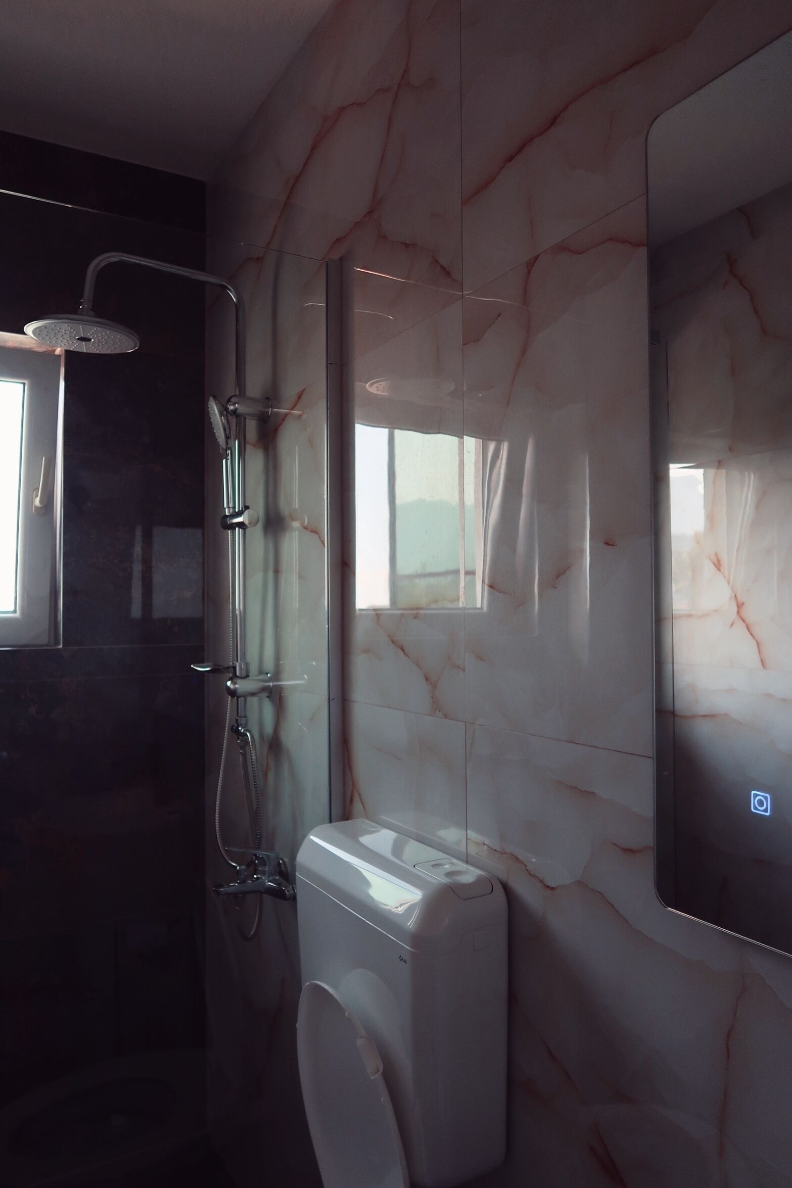 A bathroom with a marble wall, a toilet, a shower with a handheld showerhead, and a window.