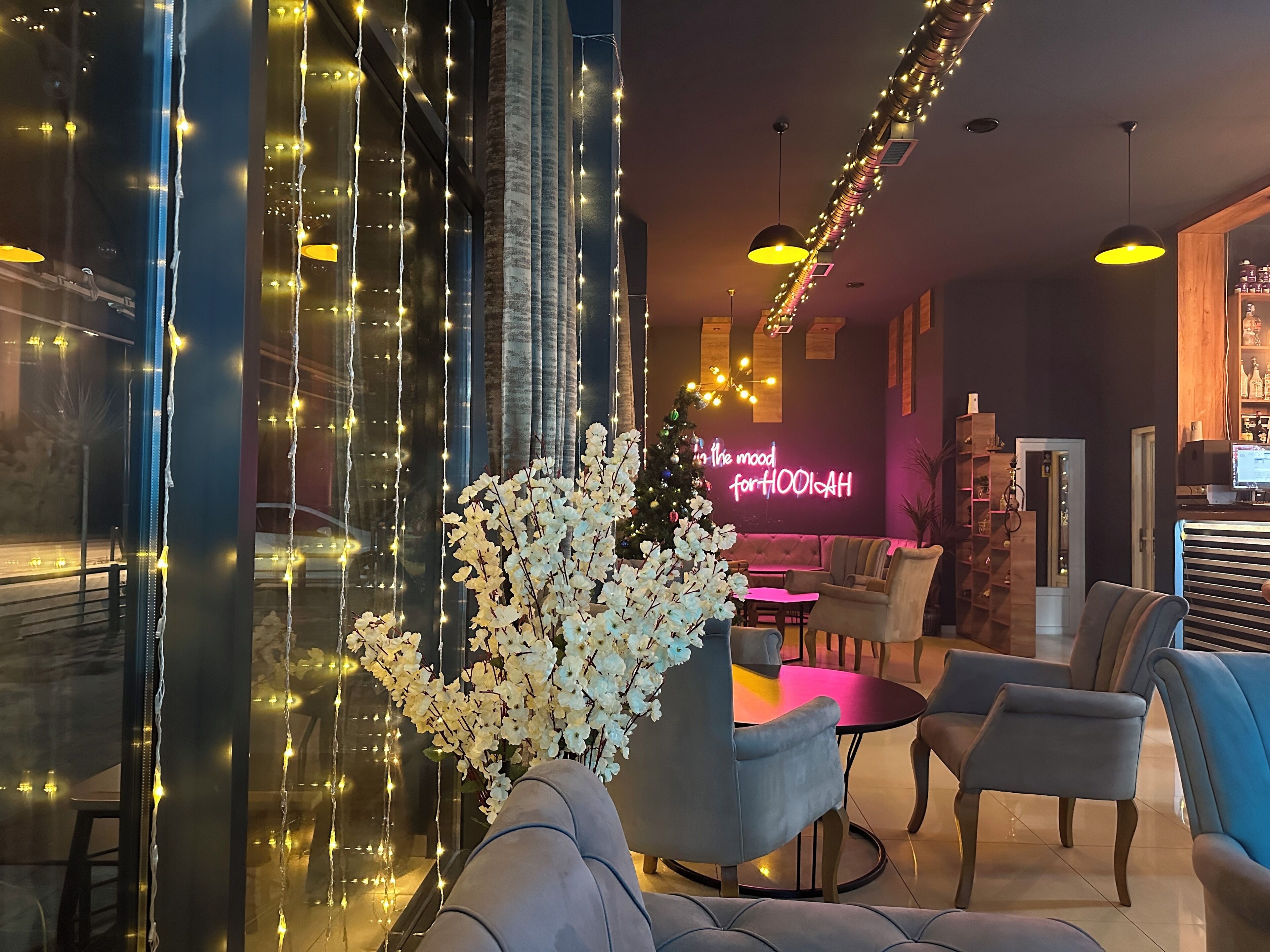A modern lounge area with a large window adorned with string lights, a seating area with sofas and tables, and a neon sign in the background.
