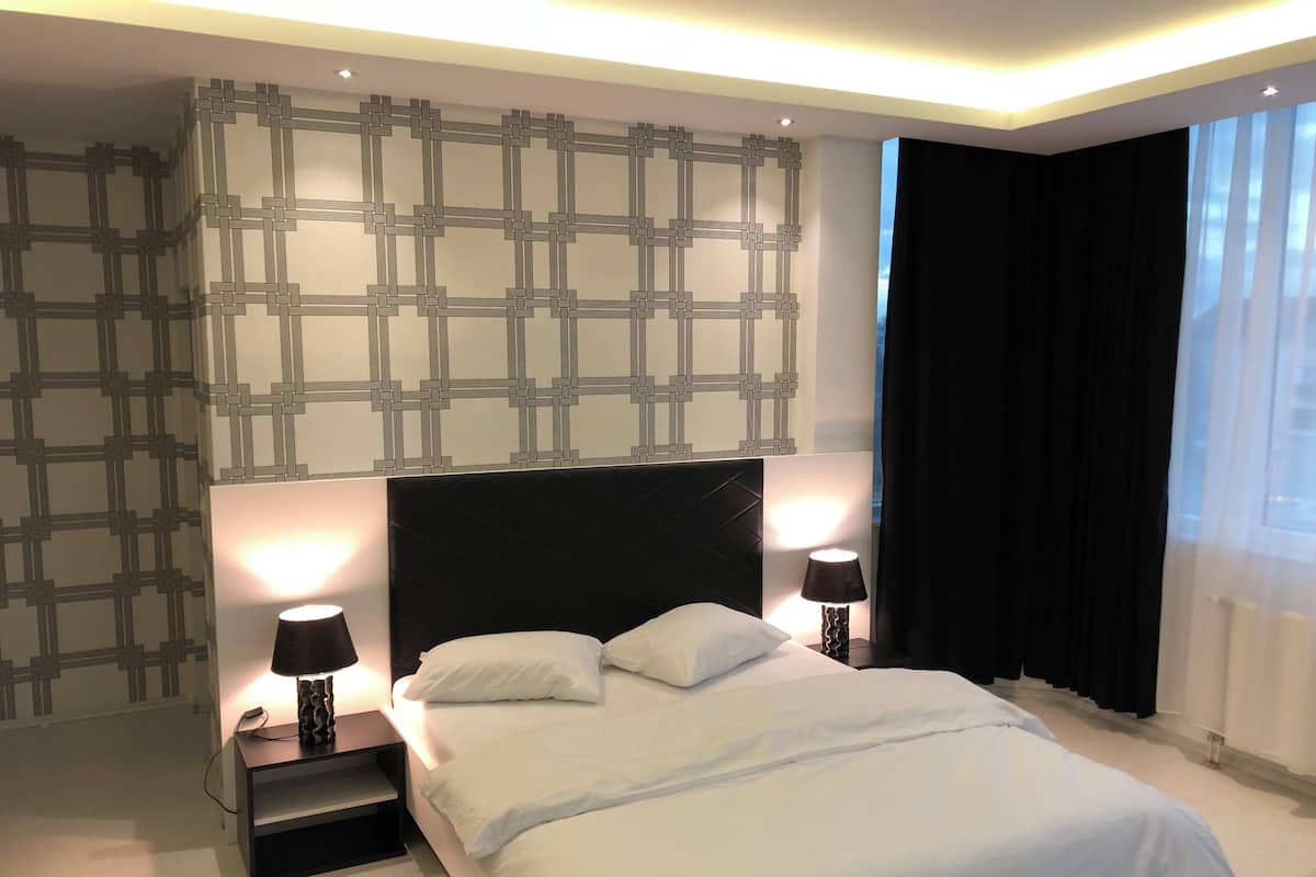 A modern hotel room with a large bed, two bedside tables with lamps, a patterned wall, and a view of the outdoors through a window with curtains.