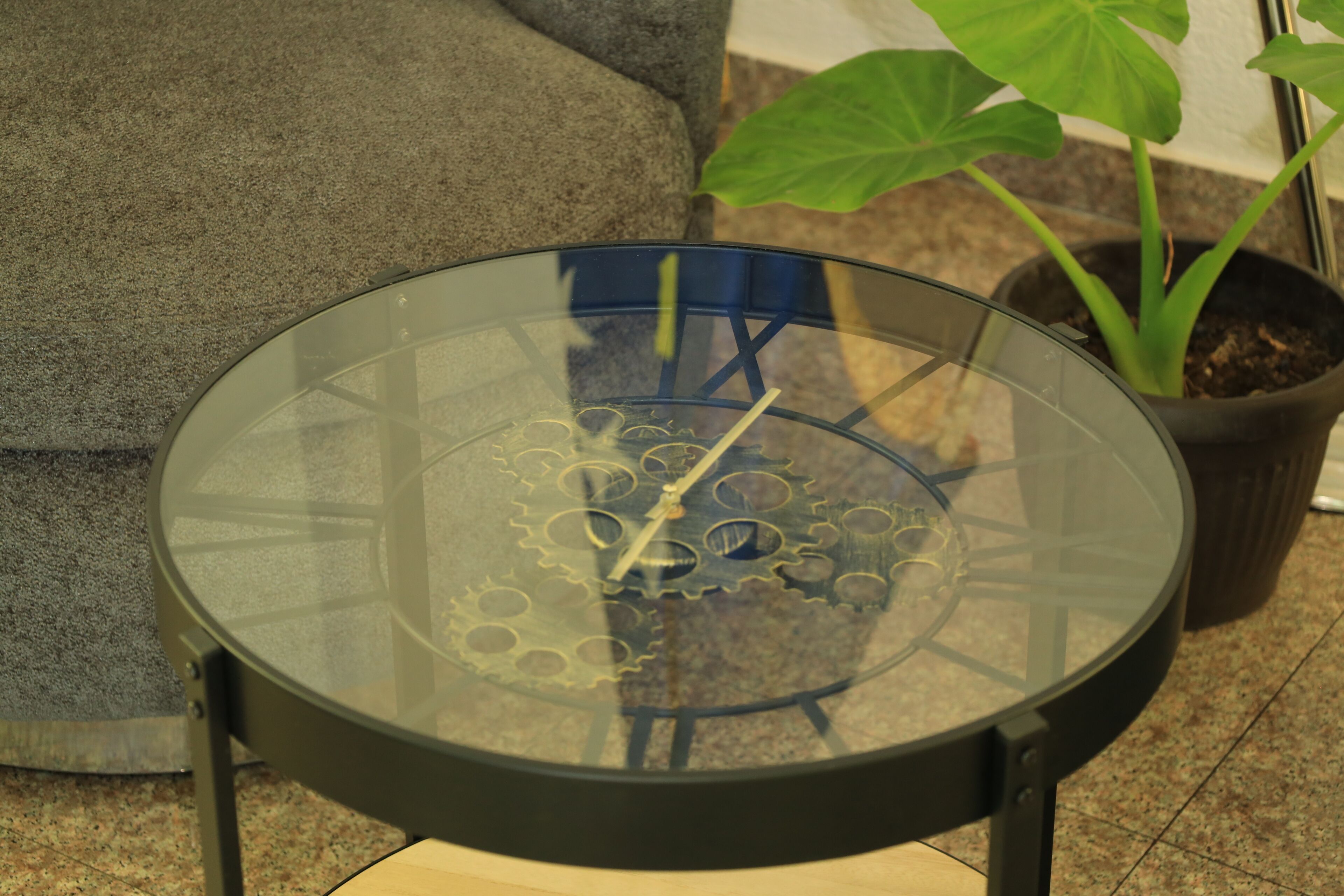 A round table with a glass top and a gear-like design, placed on a tiled floor with a potted plant nearby.