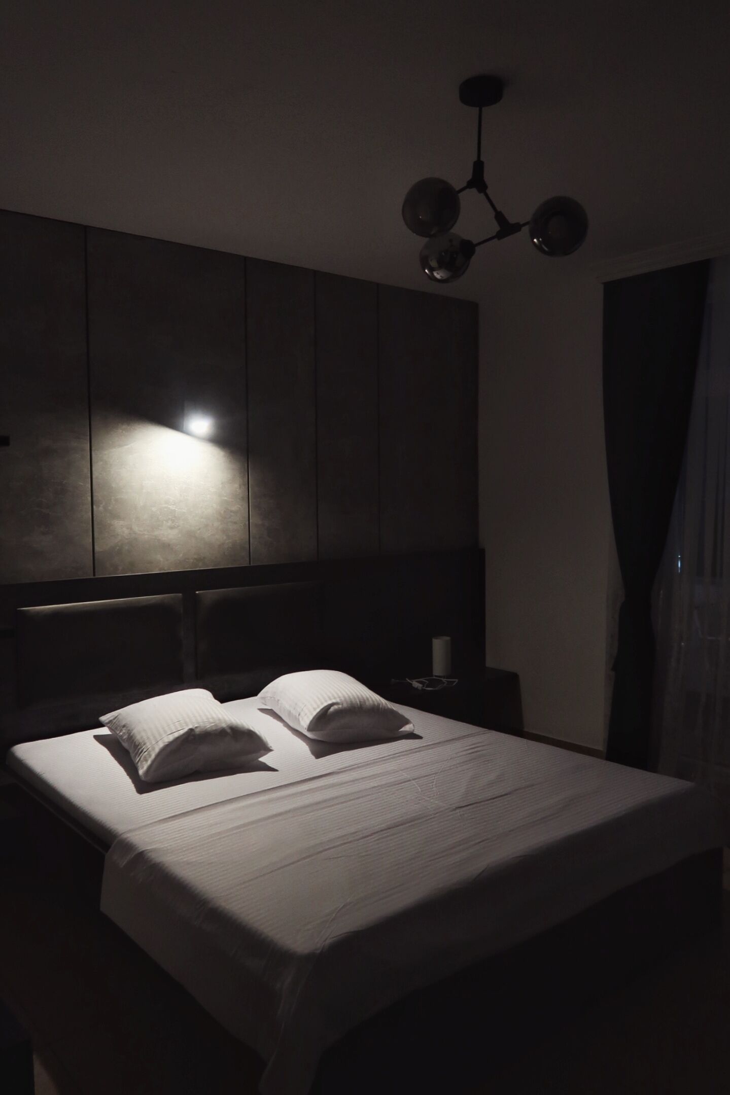 A dimly lit bedroom with a bed, a nightstand, and a wall-mounted light fixture.