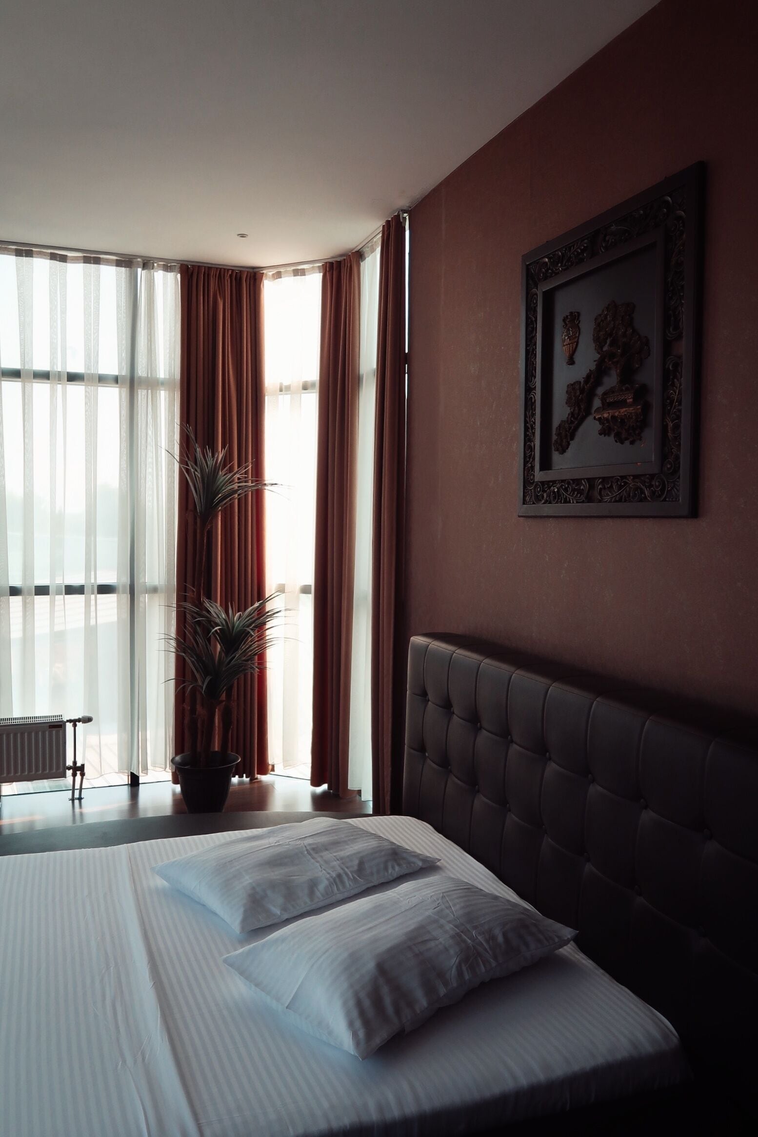 A hotel room with a large bed, a potted plant, a radiator, and a window with curtains.