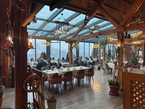 Un restaurante con techo alto, ventanas amplias, vigas de madera y lámparas colgantes.