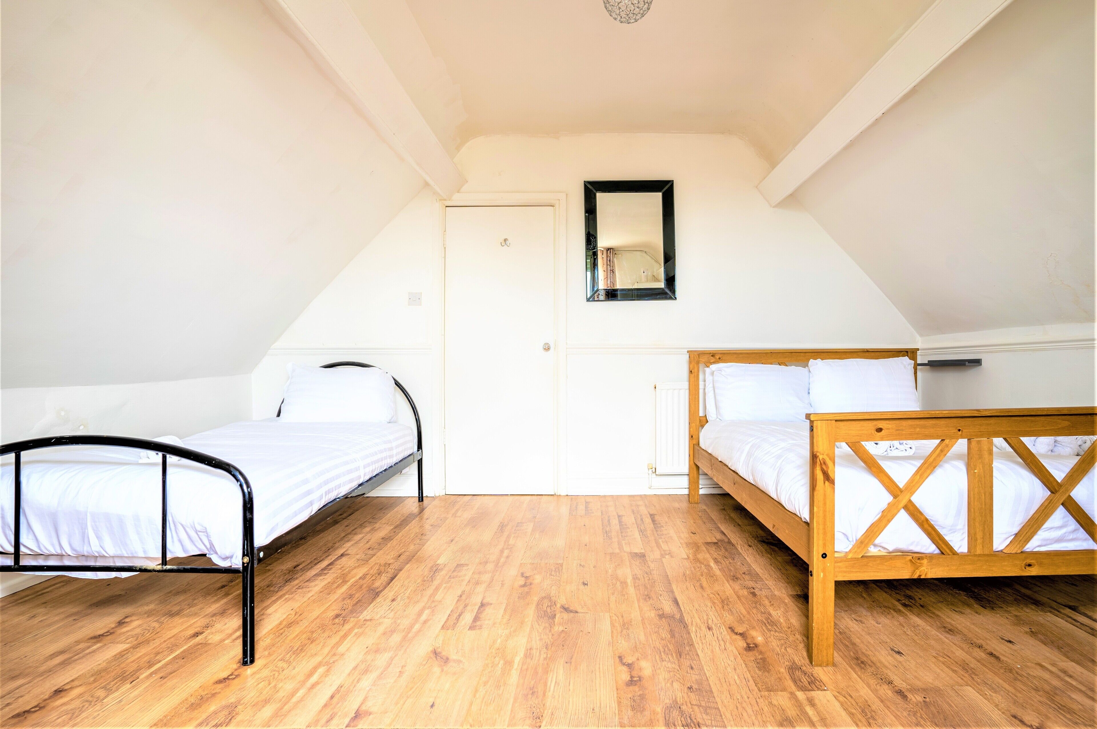A small, well-lit room with two beds, one with a wooden frame and the other with a metal frame, and a white door with a mirror above it.