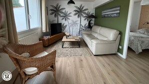 A living room with a sofa, a television on a wooden cabinet, a wicker chair, a coffee table, and a wall with palm tree wallpaper.