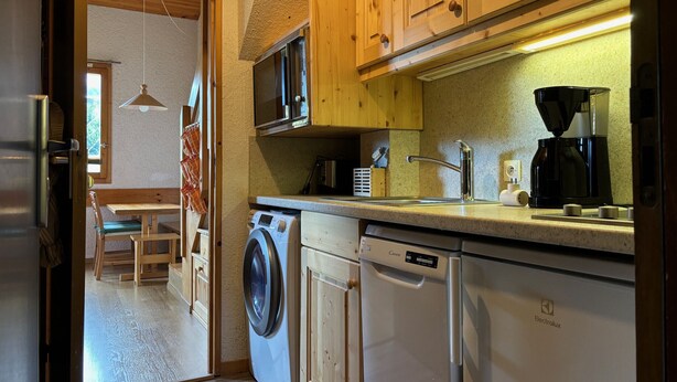 Een kleine keuken met houten kastjes, een wasmachine, een vaatwasser, een magnetron en een koffiezetapparaat.