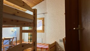 Een kamer met een houten stapelbed, een klein tafeltje en een stoel. Er is een raam met uitzicht naar buiten en een deur aan de rechterkant.