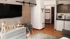 A modern living room with a grey sofa, a flat-screen TV, a small table with decorative items, and a kitchen area with cabinets and a microwave. - Modern 2-room apartment for 6 people with pool, sauna, hammam, wifi and pets allowed (Les Orres)