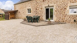 A stone building with a patio, a table with chairs, and a wooden structure to the left. - Restored 19th Century Semi-detached House in Normandy (Saint-Joseph (Manche))