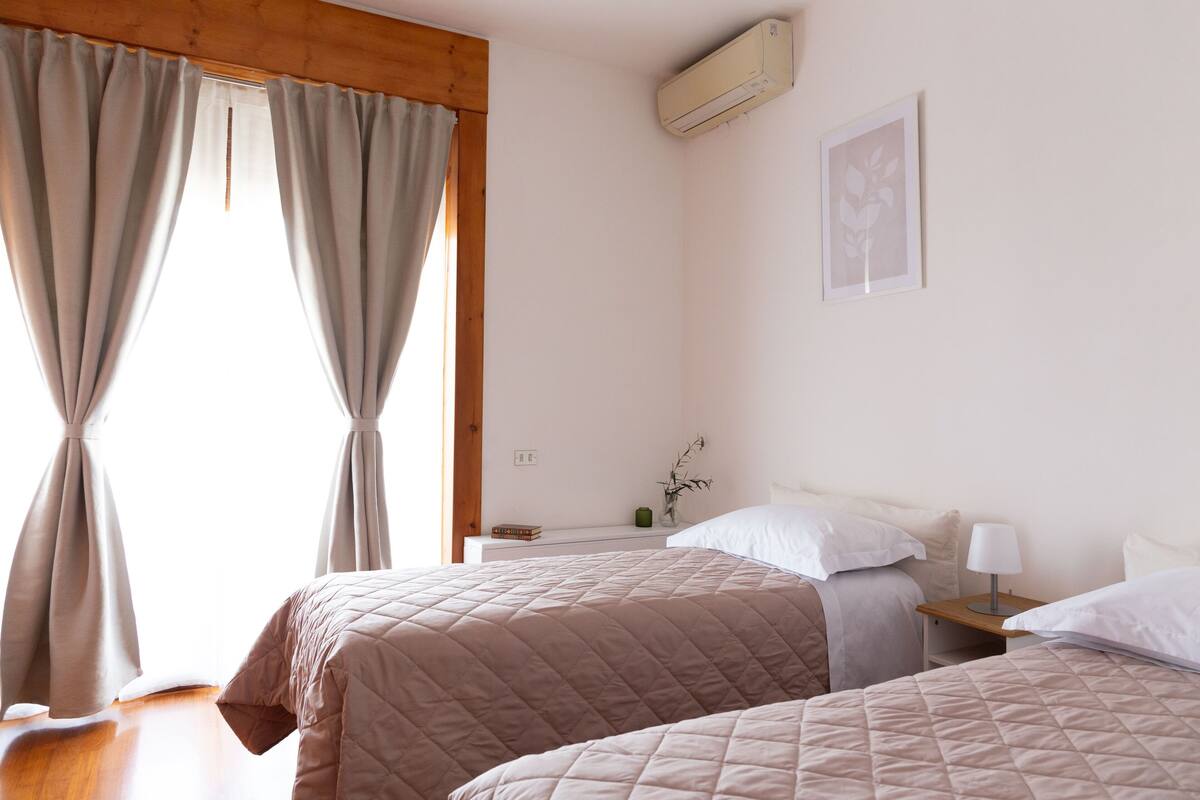 Hotelkamer met twee bedden, een houten plafond, een airconditioner, een nachtkastje met een lamp en een ingelijst kunstwerk aan de muur.