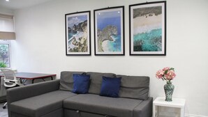 A gray sectional sofa with two blue cushions, a white side table with a vase of pink flowers, and three framed coastal landscape pictures on the wall.