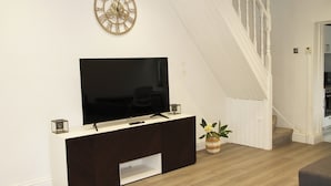A living room with a flat-screen TV on a dark wood cabinet, a decorative clock, and a staircase leading to another room.