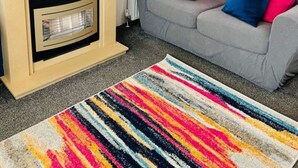 A living room with a fireplace, a sofa with colorful cushions, a round mirror, and a vibrant striped rug. - Place2bee , Caravan , Manor park , Free Wifi ,6 Berth (Hunstanton)