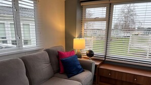 A living room with a sofa, a lamp, and a window with blinds. - Place2bee , Caravan , Manor park , Free Wifi ,6 Berth (Hunstanton)