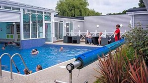 A swimming pool area with a covered section, a lounge area with seating, and a building with large windows. - Place2bee , Caravan , Manor park , Free Wifi ,6 Berth (Hunstanton)