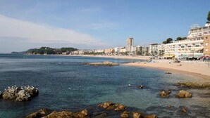 A coastal cityscape with a beach, rocky shoreline, and buildings along the waterfront. - Hotel Caleta  (Lloret de Mar)