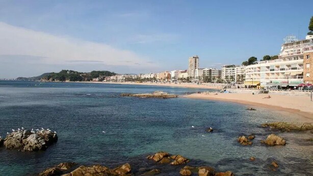 A coastal cityscape with a beach, rocky shoreline, and buildings along the waterfront. - Hotel Caleta  (Lloret de Mar)